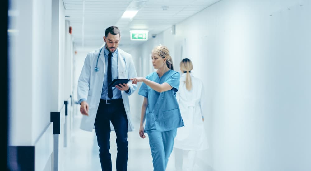 A doctor and a nurse walking through a hospital corridor with a clipboard.