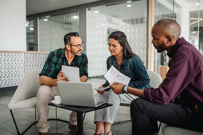 Three professionals discussing documents in a modern office lounge area.
