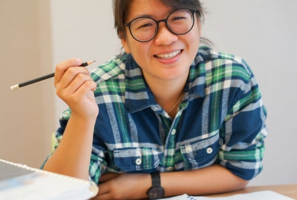 A VU Online MBA student surrounded by study materials holds up a pencil and smiles.