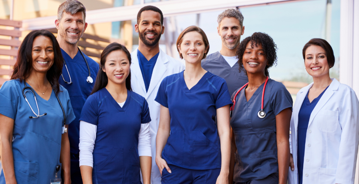 A diverse group of medical professionals smile at the camera while standing outside a building.