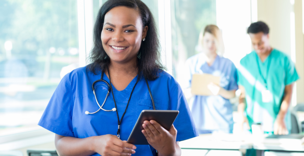 Smiling healthcare worker in blue scrubs with a stethoscope holds a tablet in a bright hospital office, with two colleagues blurred in the background near windows.