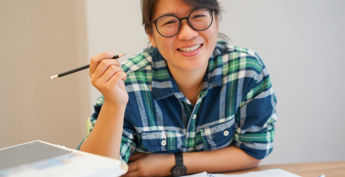 A VU Online MBA student surrounded by study materials holds up a pencil and smiles.