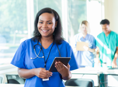 Smiling healthcare worker in blue scrubs with a stethoscope holds a tablet in a bright hospital office, with two colleagues blurred in the background near windows.