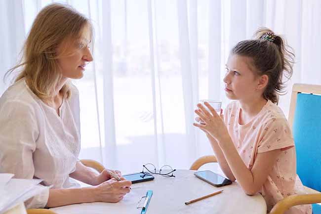 A therapist engages in a conversation with a young girl holding a glass of water.