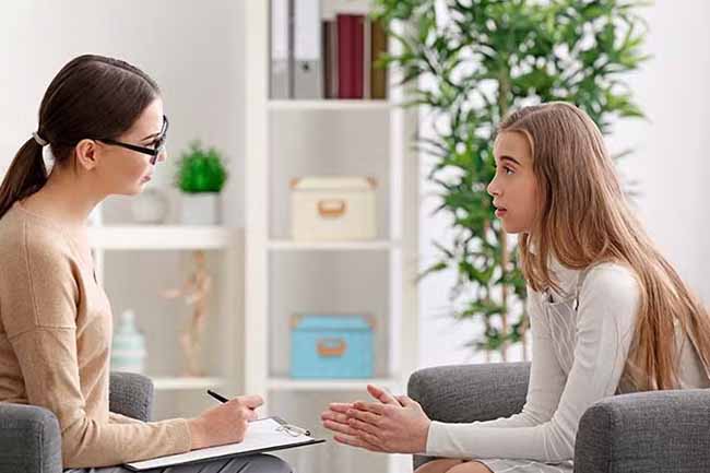 A professional counselor talks to a teenage girl in a bright office setting.